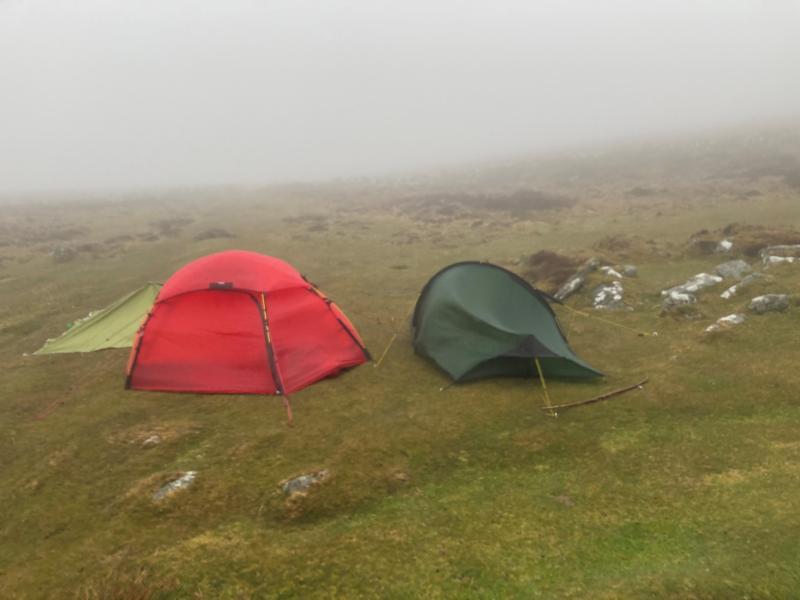 A tarp tent, red tent and a green tent are being battered by the wind on a misty moorland top