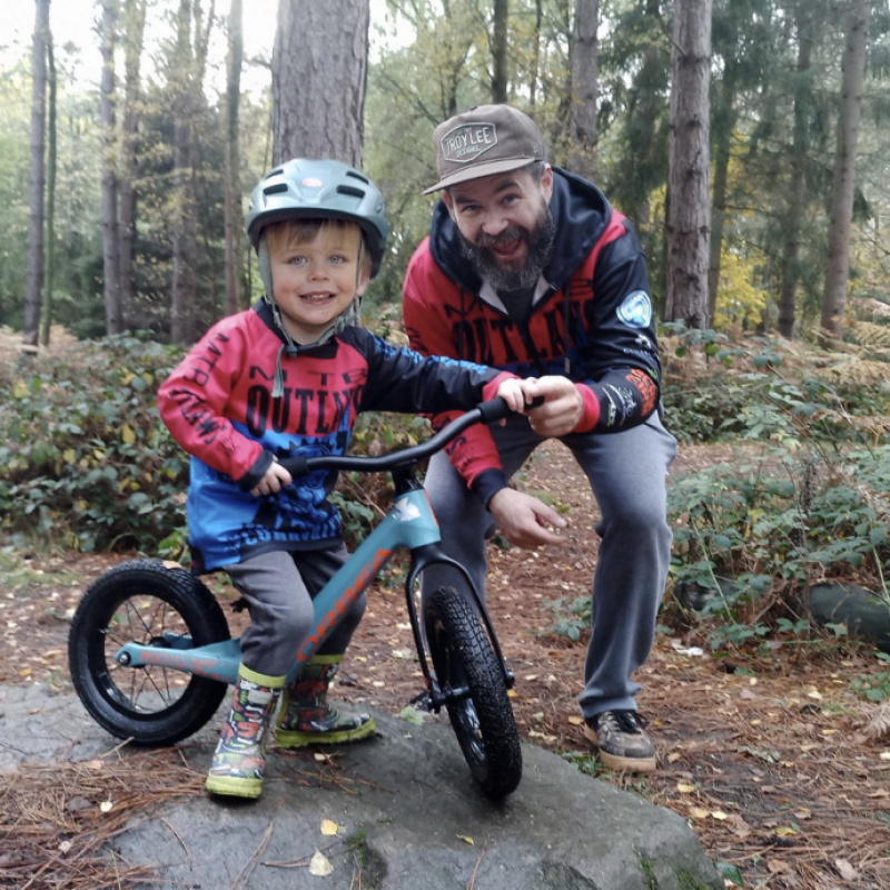A young boy is sitting on a balance bike on a flat stone in a forest. A man is with him holding onto the bike. They are both wearing MTB Outlaws cycling kit and smiling for the camera