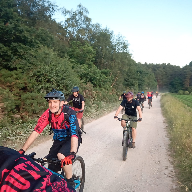 A group of cyclists on mountain bikes is riding along a gravel track next to a forest. They are all wearing MTB Outlaws red and blue kit