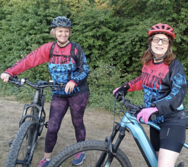 Two women are standing with their mountain bikes, one of which is an e-mountain bike, on a grassy track. They are wearing mountain biking kit and helmets.