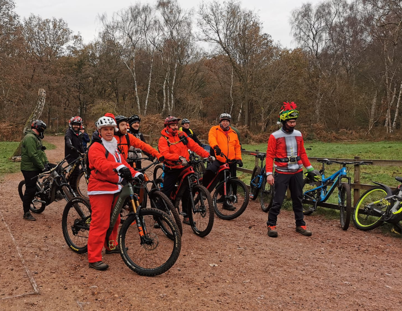 A group of cyclist on an off-road track with their mountain bikes. Most are wearing red, with some in Santa suits. It's winter.
