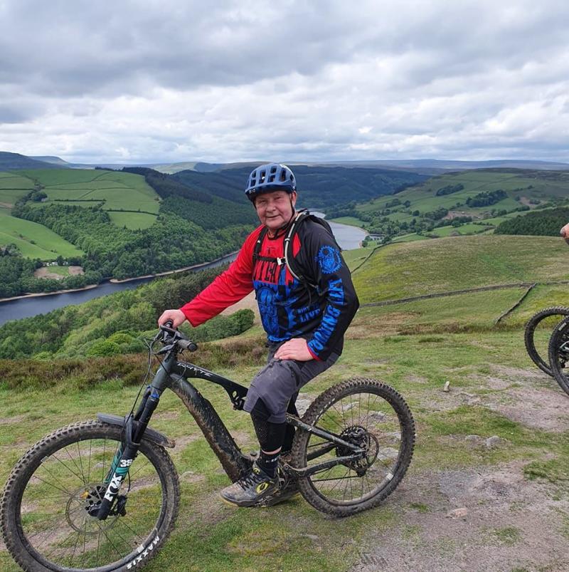 A man is sitting on a mountain bike in an off-road setting with a valley behind him with a stream running through it