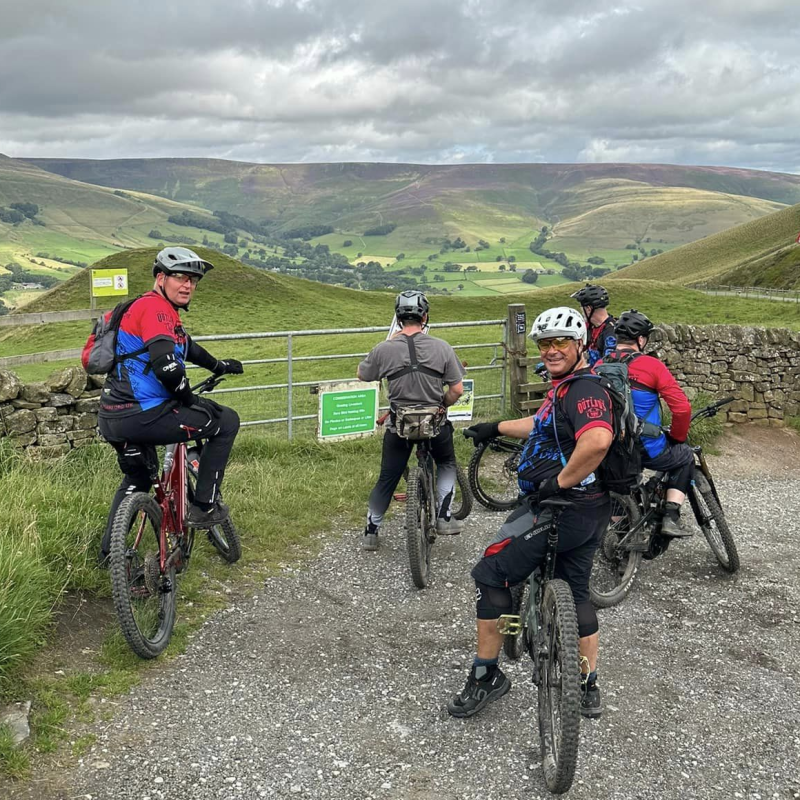A group of men on mountain bikes has stopped on a gravel track by a gate and are looking out over the undulating Peak District countryside