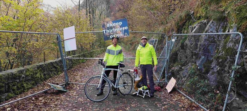 Two people are standing in front of a metal fence across a country road. They are wearing yellow hi-vis jackets. One is wearing a cycling helmet and holding a bike. The other has a cap and a loud speaker