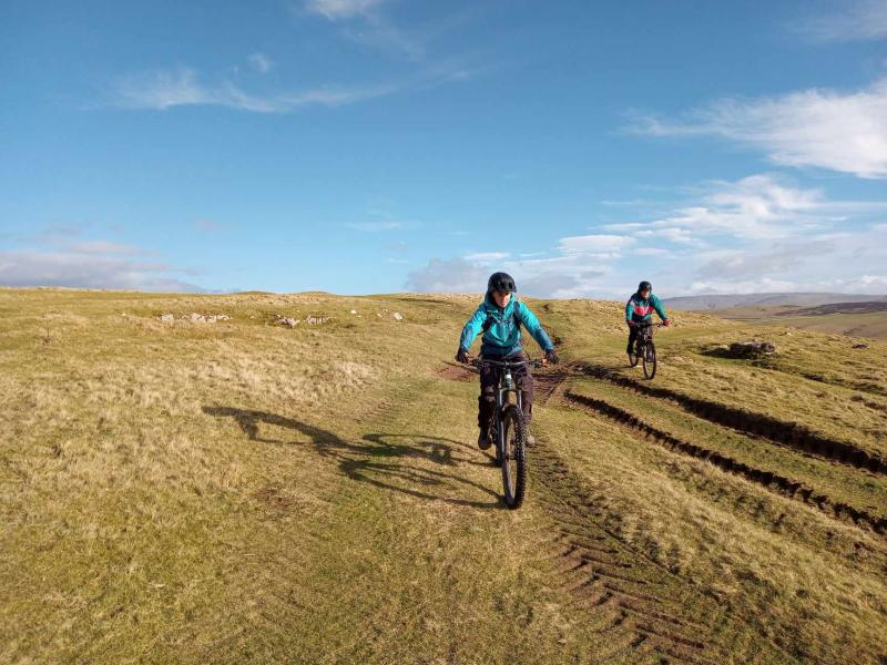 Two people are riding mountain bikes on a grassy off-road track that's rutted with car tyre tracks