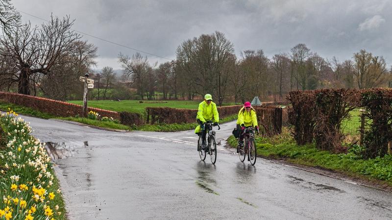 Two people are riding on a very wet village road in spring. There are daffodils growing alongside the road. The cyclists are wearing hi-vis waterproof clothing.