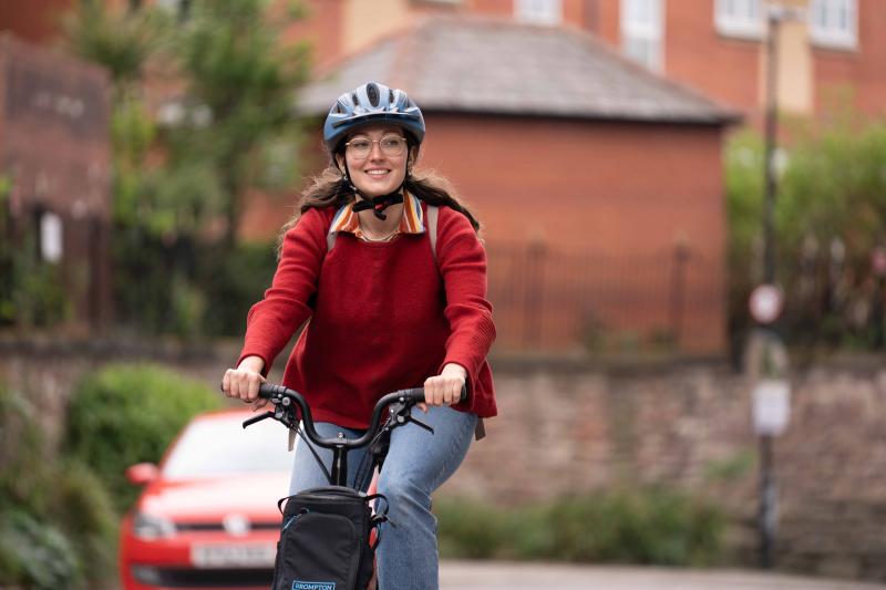 Woman wearing glasses, red jumper and blue helmet looks to the right and smiles as she pedals electric Brompton