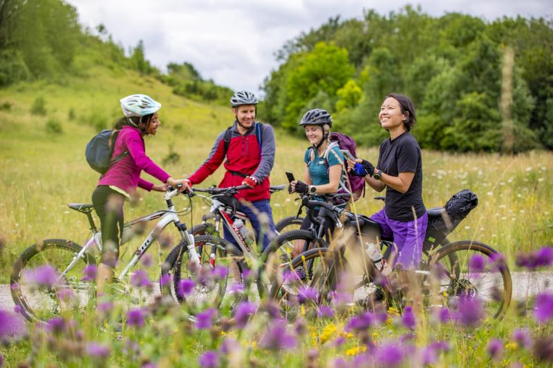 Group of four cyclists standing with their bikes during an off-road ride. Two are using their phones.