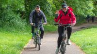 Two adults cycling on a leafy park path. The woman in front is smiling and waving, wearing a red jacket and black cap. The man behind her is also cycling, dressed in dark clothing. Lush green trees and grass surround the scene.