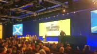 A large crowd of people sat around a stage with a panel of people sitting on it and a person standing to the right. There's a large screen that reads 'SNP 2025 annual conference'. There are also a couple of screens showing the St Andrew's Cross