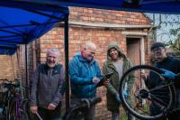 Four men smile and chat while repairing bikes outdoors under a canopy, with one holding a clipboard and another holding up a bike wheel.