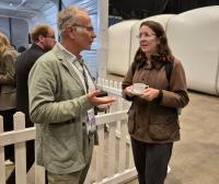 Two people, a man and a woman, are standing in front of a white picket fence. The woman is holding a cup of tea