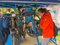 Woman in a bright orange jacket helping repair a bike under a gazebo at a Big Bike Revival event, with a second person assisting beside her.