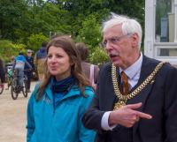 A woman in a blue Cycling UK jacket stands beside a man wearing a mayoral chain and pointing. They are outdoors at a community event, with other people and cyclists visible in the background.