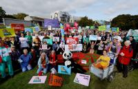 A large group of people is sitting on the grass. Some are with bikes. They are holding various flags and banners, including a bright orange one with the Cycling UK logo. In the centre is someone dressed up like a panda