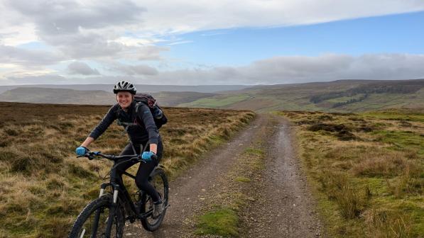 A cyclist wearing a bright green jacket and a helmet rides a mountain bike along a rugged trail through a wild and open moorland. The cloudy sky and expansive landscape create a sense of adventure and connection with nature, highlighting the joy and resilience of off-road cycling.
