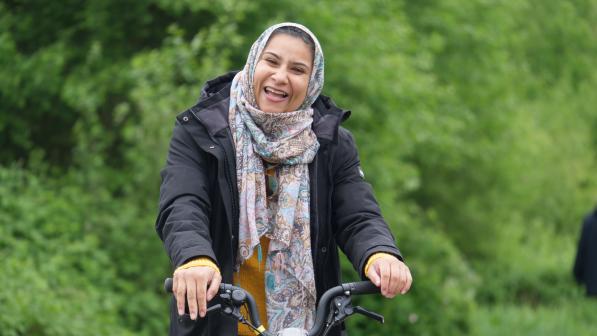 A smiling woman in a patterned headscarf and black coat stands holding a bicycle on a leafy path, enjoying a moment outdoors.