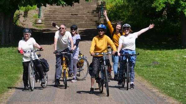 A group of cyclists, including women and men, ride together along a sunny, tree-lined path. Several riders smile and raise their arms in celebration, creating a joyful and inclusive atmosphere.