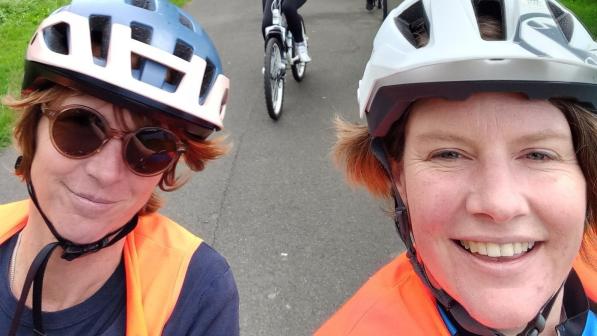 Two women in helmets and hi-vis vests smile at the camera while leading a group of young people riding bikes along a tree-lined park path behind them.
