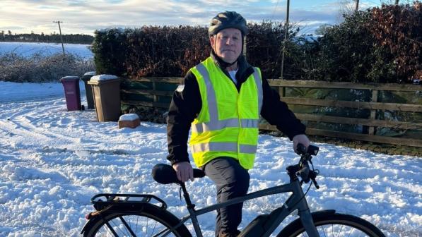 Person wearing a high-vis vest and helmet stands beside a grey e-bike on a snowy path, with hedges and a partly cloudy winter sky in the background.