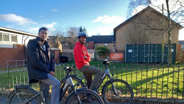 Two people sit on bicycles on a sunlit residential street, paused beside a grassy verge with low brick buildings and a fenced utility area in the background.