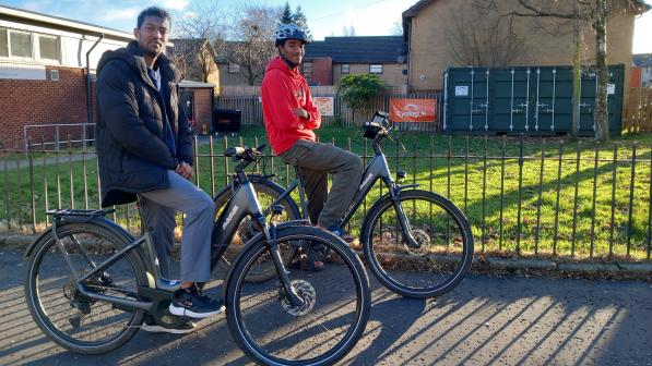 Two people sit on bicycles on a sunlit residential street, paused beside a grassy verge with low brick buildings and a fenced utility area in the background.