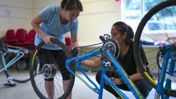 two women standing over a bike indoors. The back wheel is removed and they are working on it