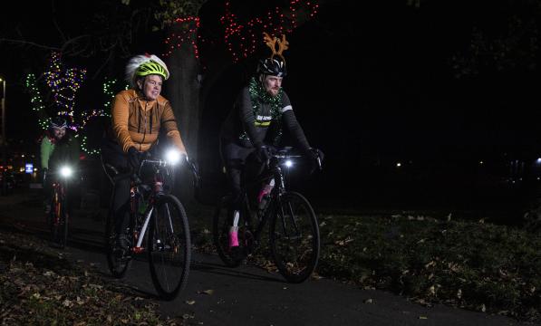 A male and female ride their bikes in the dark with front lights on. One person is wearing a pair of novelty antlers and the other is wearing a Santa hat.
