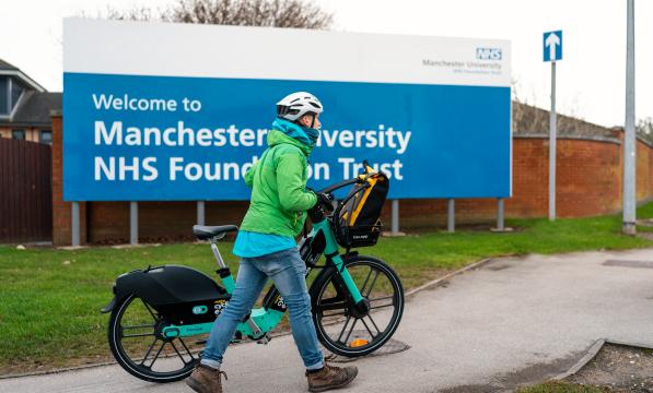 Man wearing green jacket, white helmet and blue jeans pushes TIER e-cycle past Manchester University NHS Foundation Trust sign.