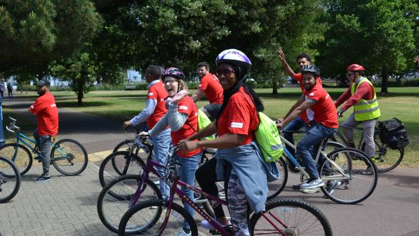 A diverse group of cyclists is cycling around a park. They are wearing matching red T-shirts and are on a variety of cycles.