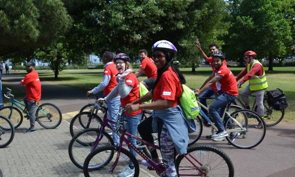 A diverse group of cyclists is cycling around a park. They are wearing matching red T-shirts and are on a variety of cycles.