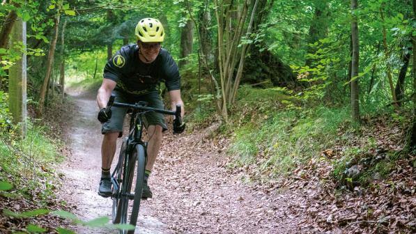 A man in cycling kit and helmet is riding a gravel bike downhill on a muddy track through a forest