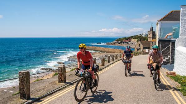 Three people are cycling along a single-lane tarmac road next to the sea