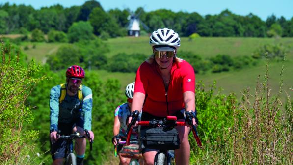 Three people on loaded gravel bikes are cycling along a grassy bush-lined track with fields and a windmill in the background