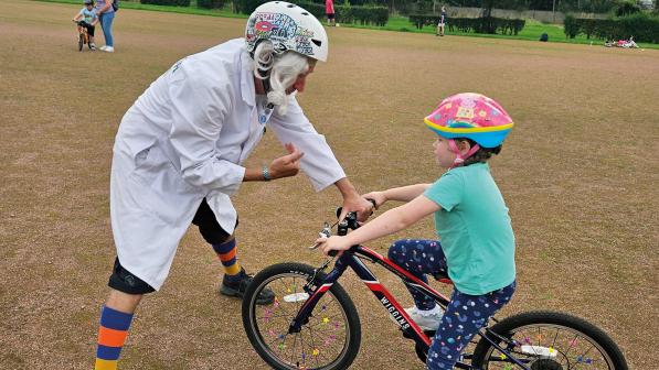A man in a white lab coat, shorts and stripey socks is holding the front of a child's bike as he teaches them to ride. 