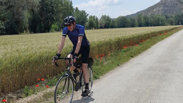 A man is cycling along a quiet tarmac road alongside a cornfield with poppies growing at the edge. There are trees and mountains in the background. He is riding a black touring bike and wearing cycling shorts and short-sleeved jersey, helmet and sunglasses