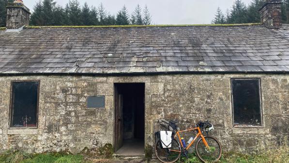 A loaded orange touring bike is leaning against an old stone hut