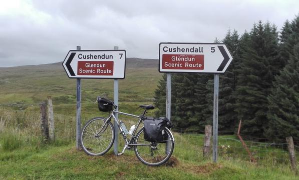 A bike is leaning up against two road signs that are pointing in opposite directions to Cushendun and Cushendall both signs also have the words Glendun Scenic Route in a brown square. In the background is a grassy hill and a line of conifers
