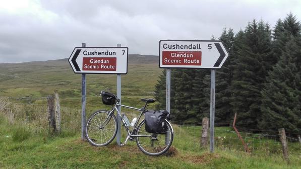 A bike is leaning up against two road signs that are pointing in opposite directions to Cushendun and Cushendall both signs also have the words Glendun Scenic Route in a brown square. In the background is a grassy hill and a line of conifers