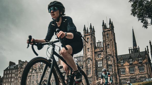 A woman in black cycling kit is riding a road bike on a road away from a castle. There is a line of cyclists behind her
