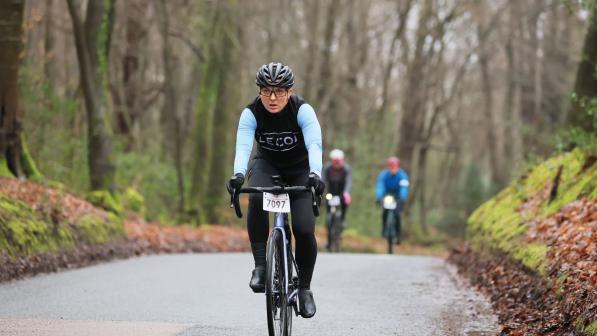 A women in cycling kit is riding a road bike with an event number on the handle bar on a country road up a hill. There are two other cyclists behind her. The road looks wet and she's wearing warm kit.