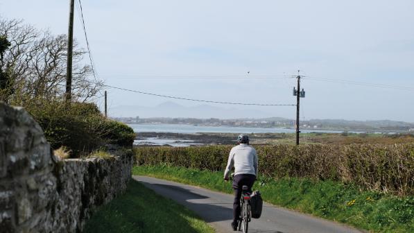 A man in a long-sleeved white top and black trousers and a cycle helmet is riding a loaded hybrid bike along a single-track road line with a dry stone wall and a hedge. A large lough is in the background