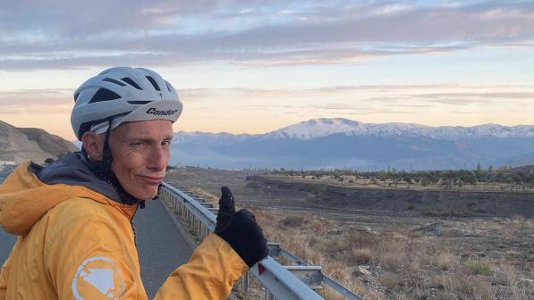 A man in cycling helmet and yellow waterproof is standing at the road side, giving a thumbs-up and looking out over a valley and mountains