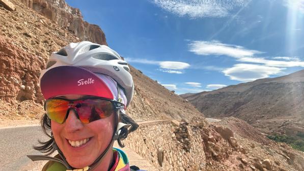 A selfie of a woman in cycle helmet, cap and sun glasses, behind her is a winding mountain road and mountains
