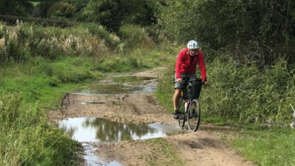 A man in red top and shorts and a cycle helmet is riding a loaded flatbar bike around a puddle on an off-road track