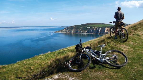 One bike lying on the ground with another being sat on as a man looks out from a clip top over the sea