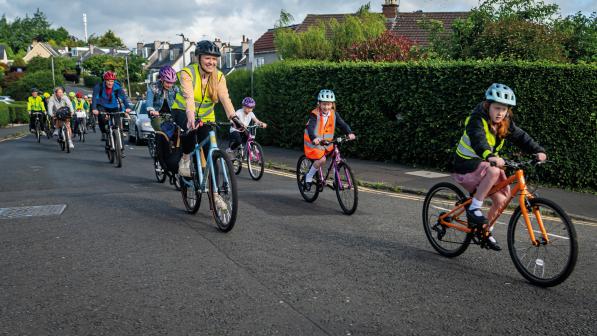 A big group of adults and children are cycling on an urban road. Many are wearing hi-vis jackets and cycle helmets. They'e smiling.