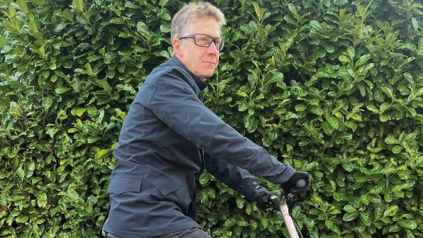 A man in jeans and a blue waterproof cycling jacket is standing astride a silver folding bike on the edge of the curb