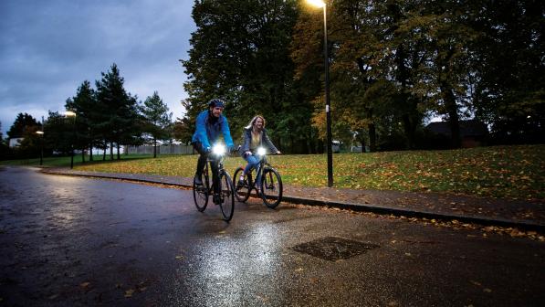 Two people are riding along a lit street in the evening. It's autumn and the road is wet and there are fallen leaves on the ground. They are wearing warm clothing and their bikes have lights on them