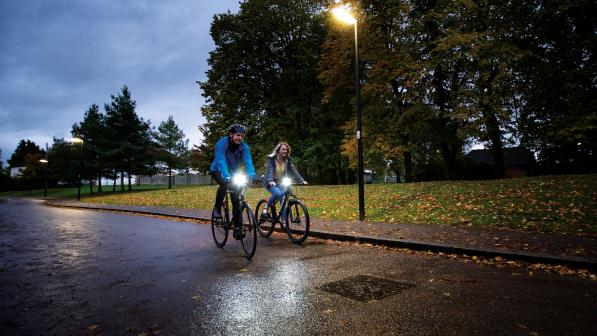 Two people are riding along a lit street in the evening. It's autumn and the road is wet and there are fallen leaves on the ground. They are wearing warm clothing and their bikes have lights on them
